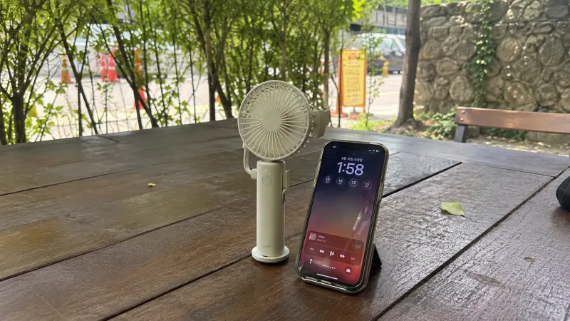 야외 그늘진 테이블에서 휴대용 쿨링팬과 함께 놓인 아이폰 모습